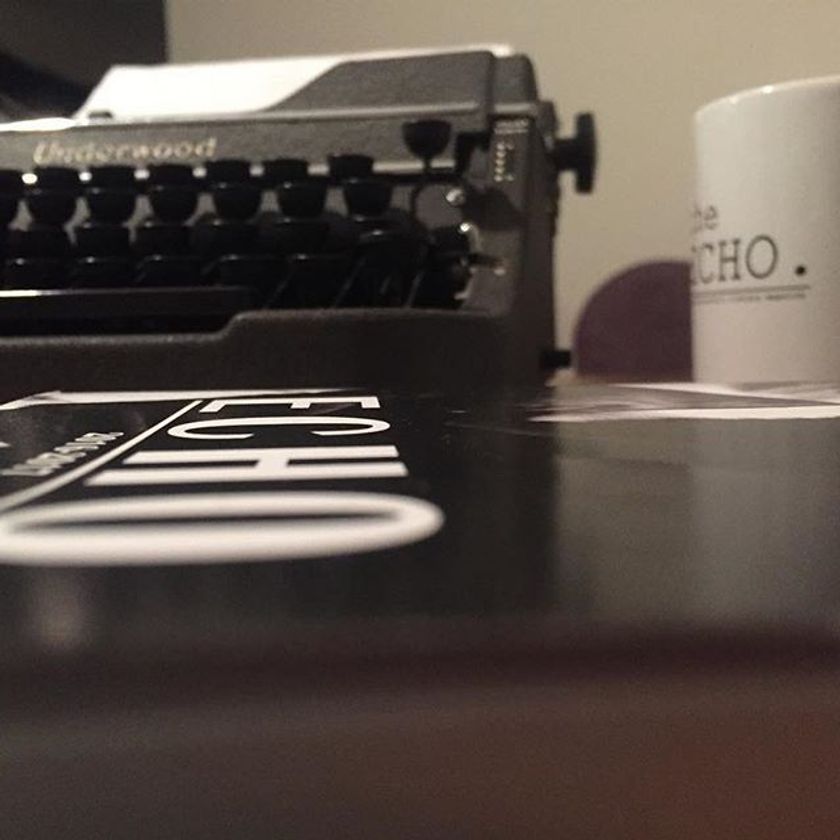 A close side view of a table with on old copy of The Echo, an Underwood typewriter, and a mug with The Echo's logo.