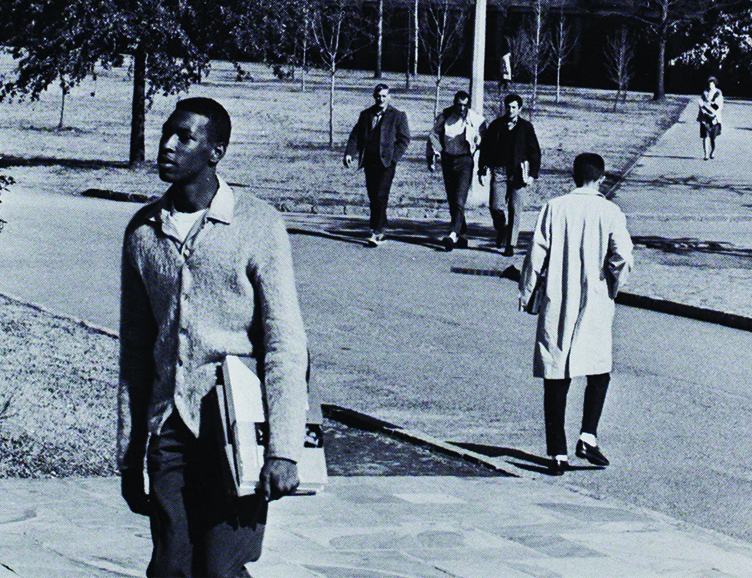 Black-and-white photo of Joseph Vaughn walking across campus. Other students are in the background.